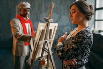 Portrait painter at the easel draws female model. Male artist standing at his workplace, creative master works in workshop
