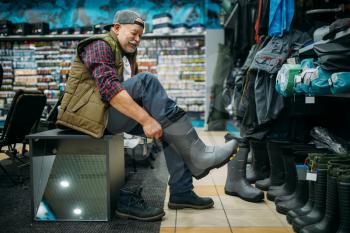 Fisherman tries on rubber boots in fishing shop. Equipment and tools for fish catching and hunting, accessory choice on showcase in store