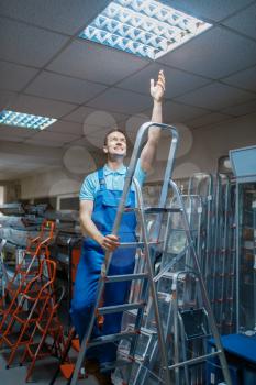 Male worker in uniform standing on stepladder in tool store. Department with ladders, choice of equipment in hardware shop, instrument supermarket