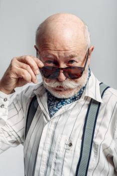 Bearded elderly man with mustache poses in sunglasses, grey background. Mature senior looking at camera