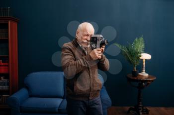 Elderly man poses with old film camera in home office. Bearded mature senior in living room, old age businessman