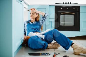 Tired female plumber in uniform installing drain pipe in the kitchen. Handywoman with toolbag repair sink, sanitary equipment service at home