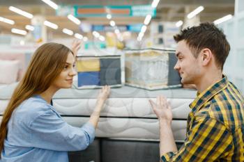Young love couple choosing mattress for the bed in furniture store showroom. Man and woman looking samples for bedroom in shop, husband and wife buys goods for modern home interior