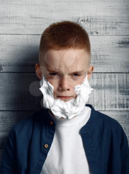 Serious little boy with shaving foam on his face in studio. Kid isolated on wooden background, child emotion, schoolboy photo session
