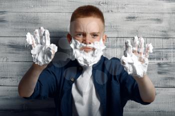 Little boy with shaving foam on his face and hands in studio. Kid isolated on wooden background, child emotion, schoolboy photo session