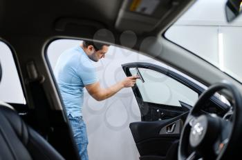 Male wrapper installs car tinting, tuning service. Worker applying vinyl tint on vehicle window in garage, tinted automobile glass