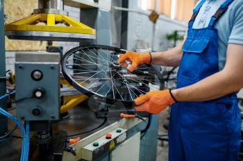 Mechanic in uniform holds bicycle wheel near machine tool on factory. Bike rims and spokes assembly in workshop, cycle parts installation, modern technology