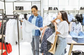 Two girls choosing trousers in clothing store. Women shopping in fashion boutique, shopaholics, shoppers looking garment on hangers