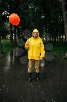 Sad man with balloon walking in summer park in rainy day. Male person in rain cape and rubber boots, wet weather in alley, loneliness