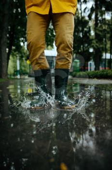 Male person in rain cape and rubber boots jumping in puddles, wet weather in alley. Man poses in summer park, rainy day. Water protection, drops
