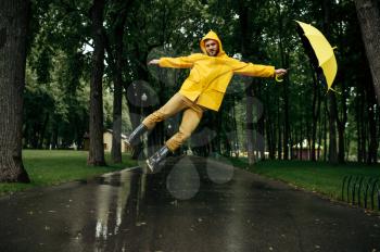 Man flying with umbrella in summer park in windy rainy day. Male person in rain cape and rubber boots, wet weather in alley