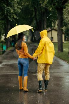 Love couple with umbrella walking in summer park in rainy day. Man and woman in rubber boots on walking path, wet weather in alley