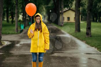 Sad woman with balloon walking in summer park in rainy day. Female person in rain cape and rubber boots, wet weather in alley, loneliness