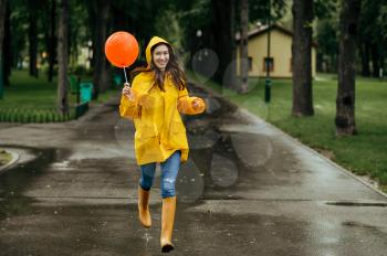 Happy woman runs with balloon in summer park in rainy day. Female person in rain cape and rubber boots, wet weather in alley, loneliness