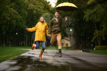 Happy love couple jumps like a children in park, summer rainy day. Man and woman under umbrella in rain, romantic date on walking path, wet weather in alley