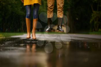 Love couple jumps in a puddle in park, summer rainy day. Man and woman under umbrella in rain, romantic date on walking path, wet weather in alley