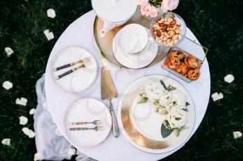 Table setting, ceramic teapot, teacups, cake and flowers closeup, side view, nobody. Luxury silverware on white tablecloth, tableware outdoors