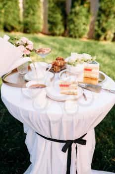 Table setting, tea party, drink is poured from ceramic teapot. Luxury silverware on white tablecloth, tableware outdoors. Celebration on summer meadow