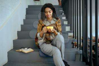 Smiling female student with coffee sitting on on the steps in library cafe. Woman with cup, opened book and glasses on background, education and knowledge. Girl studying in campus