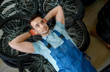 Male worker lying on stack of bicycle wheels on factory. Bike rims assembly line in workshop, cycle parts installation, modern technology