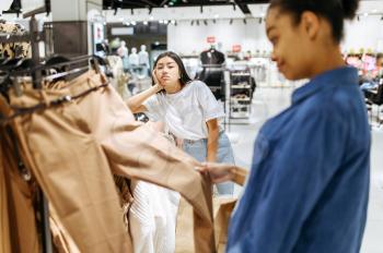 Two girls choosing trousers in clothing store. Women shopping in fashion boutique, shopaholics, shoppers looking garment on hangers