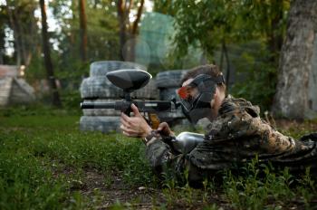 Male paintball warrior with gun shoots lying down on the grass, playground in the forest on background. Extreme sport with pneumatic weapon and paint bullets or markers, military team game outdoors