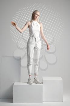 Futuristic young woman in white clothes stands on cube, light grey background. Female person in virtual reality style, future technology, futurism concept, cyber or robot theme