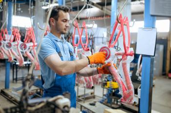 Bicycle factory, worker holds pink kid's bike frame. Male mechanic in uniform installs cycle parts, assembly line in workshop, industrial manufacturing