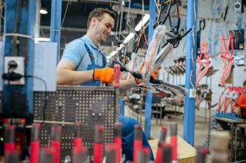 Bicycle factory, worker holds pink kid's bike. Male mechanic in uniform installs cycle parts, assembly line in workshop, industrial manufacturing