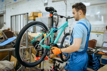 Bicycle factory, worker holds mountain bike. Male mechanic in uniform installs cycle parts, assembly line in workshop, industrial manufacturing