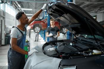 Two male workers inspects engine, car service. Vehicle repairing garage, men in uniform, automobile station interior on background. Professional auto diagnostic