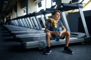 Youngster with water sitting on treadmill in gym, running machine. Boy on training in sport club, health care and healthy lifestyle, schoolboy on workout