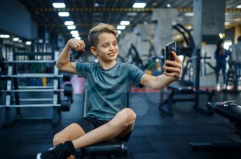 Boy with phone makes selfie on bench in gym. Youngster on training in sport club, healthcare and healthy lifestyle, schoolboy on workout