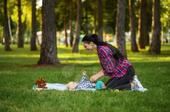 Mother play with her little son on the grass in summer park. Mom with male kid leisures on lawn, picnic with child on plaid in the forest, family on vacations