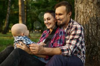 Mother, father and little baby sitting under the tree in summer park. Mom and dad with male kid on lawn, picnic with child on plaid in the forest, family happiness