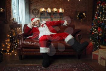 Bad Santa claus with hangover sitting on couch. Unhealthy lifestyle, bearded man in christmas costume, new year and alcoholism