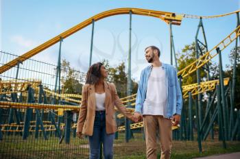 Love couple at the roller coaster in amusement park, attraction on background. Man and woman relax outdoors. Family leisures in summertime, entertainment theme
