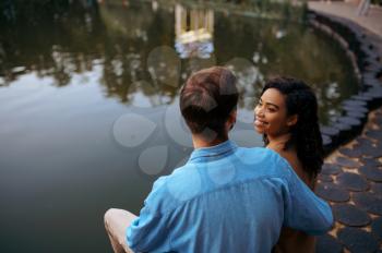 Love couple embracing at the pond in summer park. Man and woman relax outdoors, green lawn on background. Family hugging near the lake in summertime, weekend outdoors