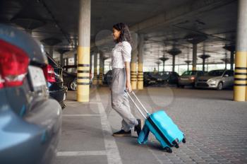 Young woman with suitcase in car parking. Female traveler with luggage in vehicle park lot, passenger with bag. Girl with baggage near automobile