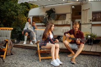 Friends playing on guitar on picnic at camping in the forest. Youth having summer adventure on rv, camping-car on background. Two couples leisures, travelling with trailer