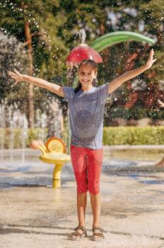 Little funny girl in splashes on water playground in summer park. Child leisures in aquapark, aquatic adventure, female kid having fun in a fountain, waterpark