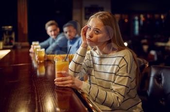Bored girl drinks coctail at the counter in bar. Group of people relax in pub, night lifestyle, female person in nightclub