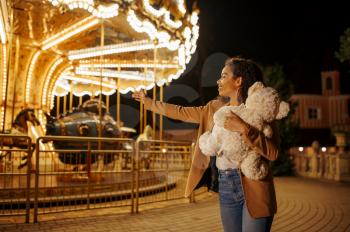 Young woman with soft toy near the carousel in night amusement park. Love couple relax outdoors, roundabout attraction with lights on background. Family leisures in summertime, entertainment theme