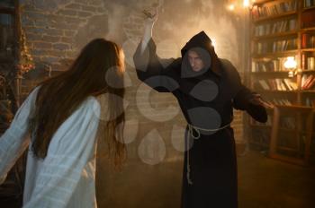 Male exorcist in black hood casting out satan from crazy woman. Exorcism, mystery paranormal ritual, dark religion, night horror, potions on shelf on background