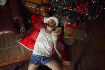 Bad shameless Santa claus in underpants lying under christmas tree, nasty party, humor. Unhealthy lifestyle, bearded man in holiday costume, new year and alcoholism