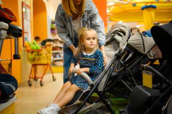 Mother and pretty little girl choosing baby stroller in store. Mom and adorable daughter near the showcase in children's shop, happy childhood, family makes a purchase in market