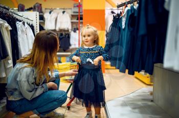 Mother and little baby choosing dress in kid's store. Mom and adorable girl near the showcase in children's shop, happy childhood, family makes a purchase in kid's market