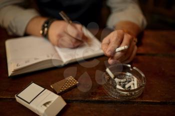 Man smokes cigarette and writes in notebook, wooden tabler on background. Tobacco smoking culture, specific flavor. Male smoker