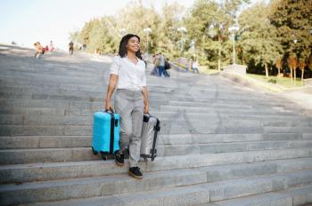 Woman carries two suitcases by the stairs in summer park. Female traveler with luggage leisures outdoors, passenger with bag resting in nature. Girl with baggage relax on city alley