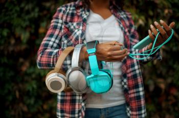 Female music fan holds headphones in summer park. Woman walking outdoors, girl in earphones, green bushes on background